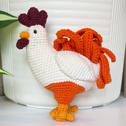 A crocheted rooster figurine with a white body, orange legs and tail feathers, and a dark burgundy comb and wattle, standing in front of a white ribbed cylindrical container.