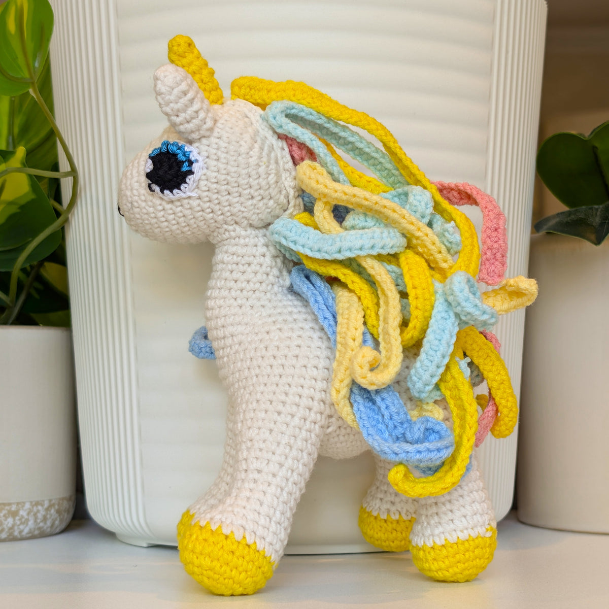 A handmade crocheted unicorn toy with a white body, yellow horn and hooves, and a multicolored mane of yellow, light blue, and pink yarn, standing in front of a white ribbed ceramic pot.