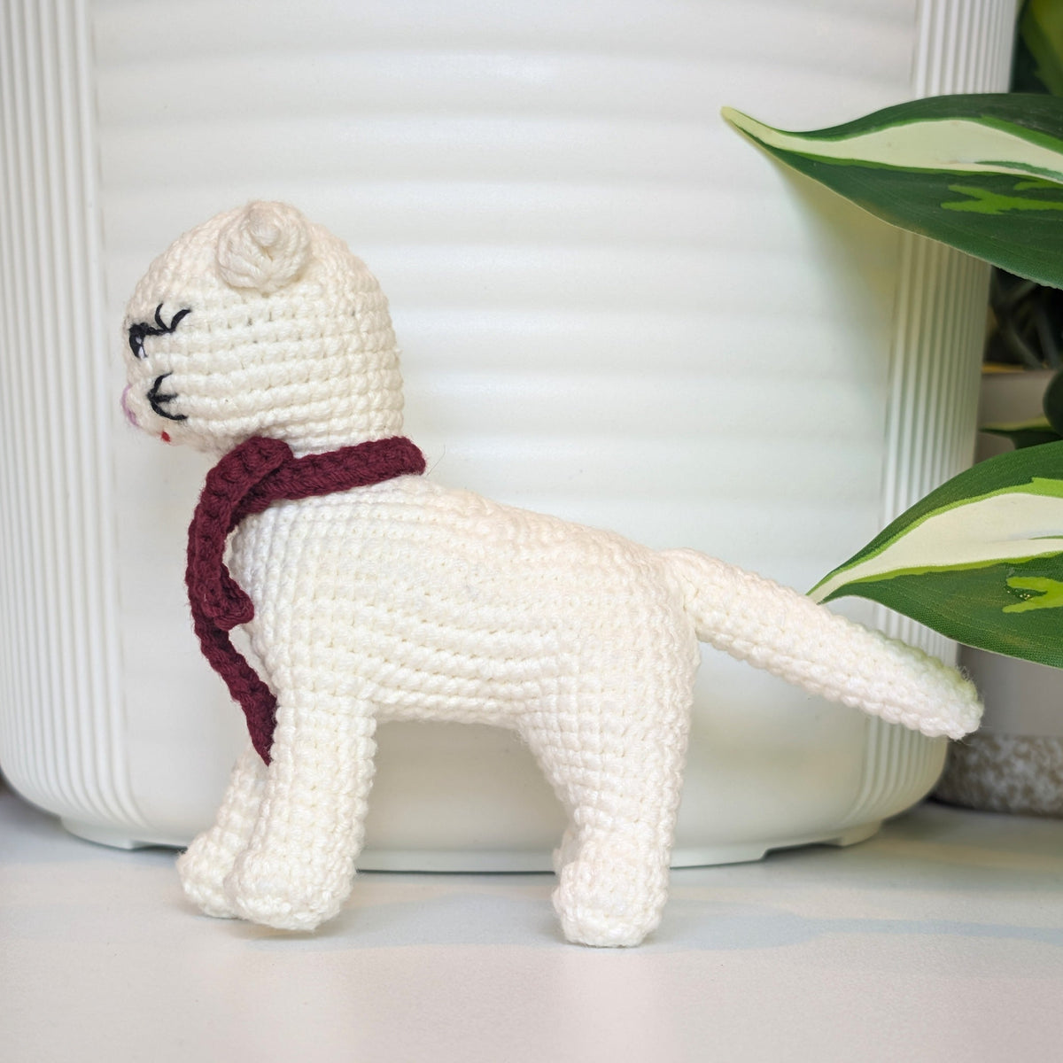 A crocheted white cat toy wearing a burgundy scarf, standing in profile in front of a white ribbed planter with green leaves on the left.